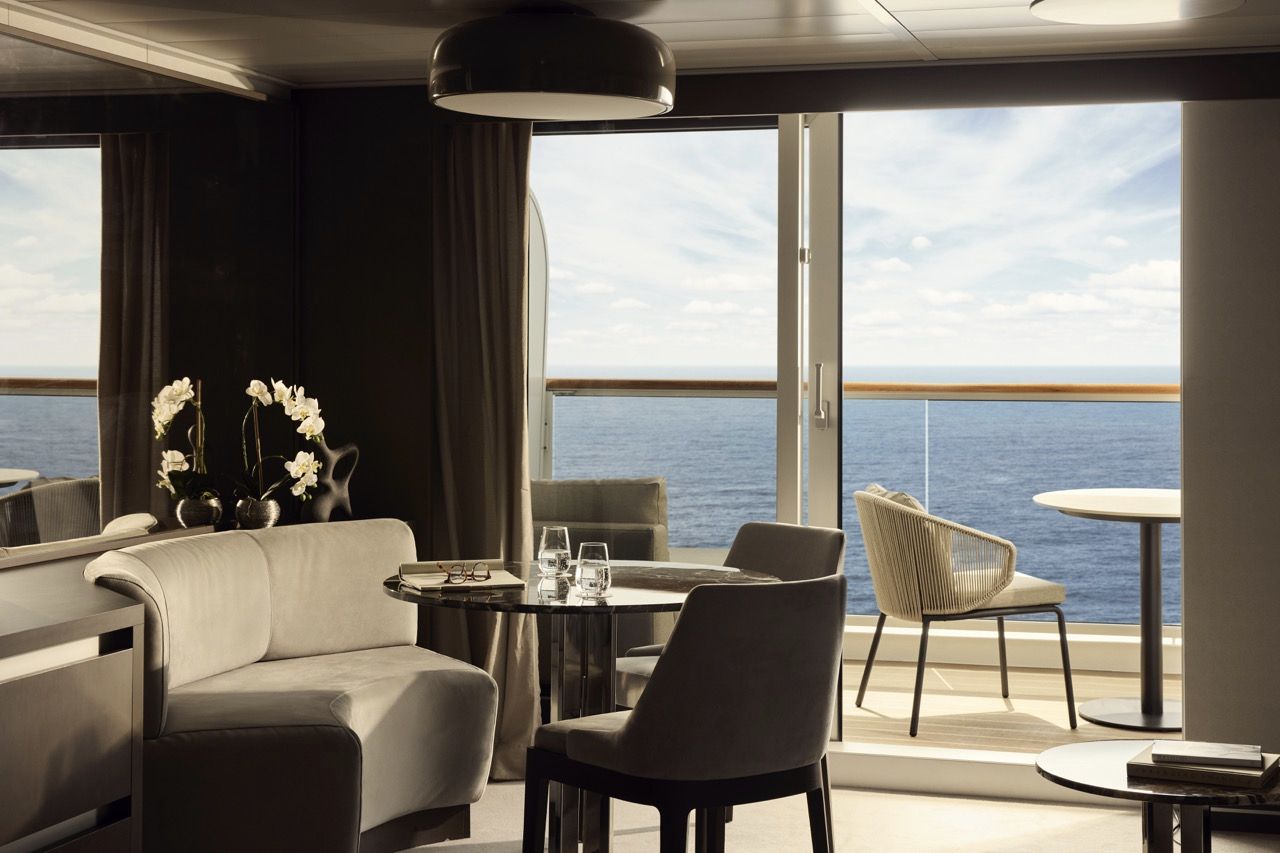 Luxuri&ouml;ser Wohnbereich eines Penthouses an Bord von Explora Journeys mit hellem Designer-Sofa, rundem Tisch und ge&ouml;ffneter Glasfront, die auf eine private Terrasse mit Sitzgelegenheiten und freiem Blick auf das blaue Meer f&uuml;hrt.