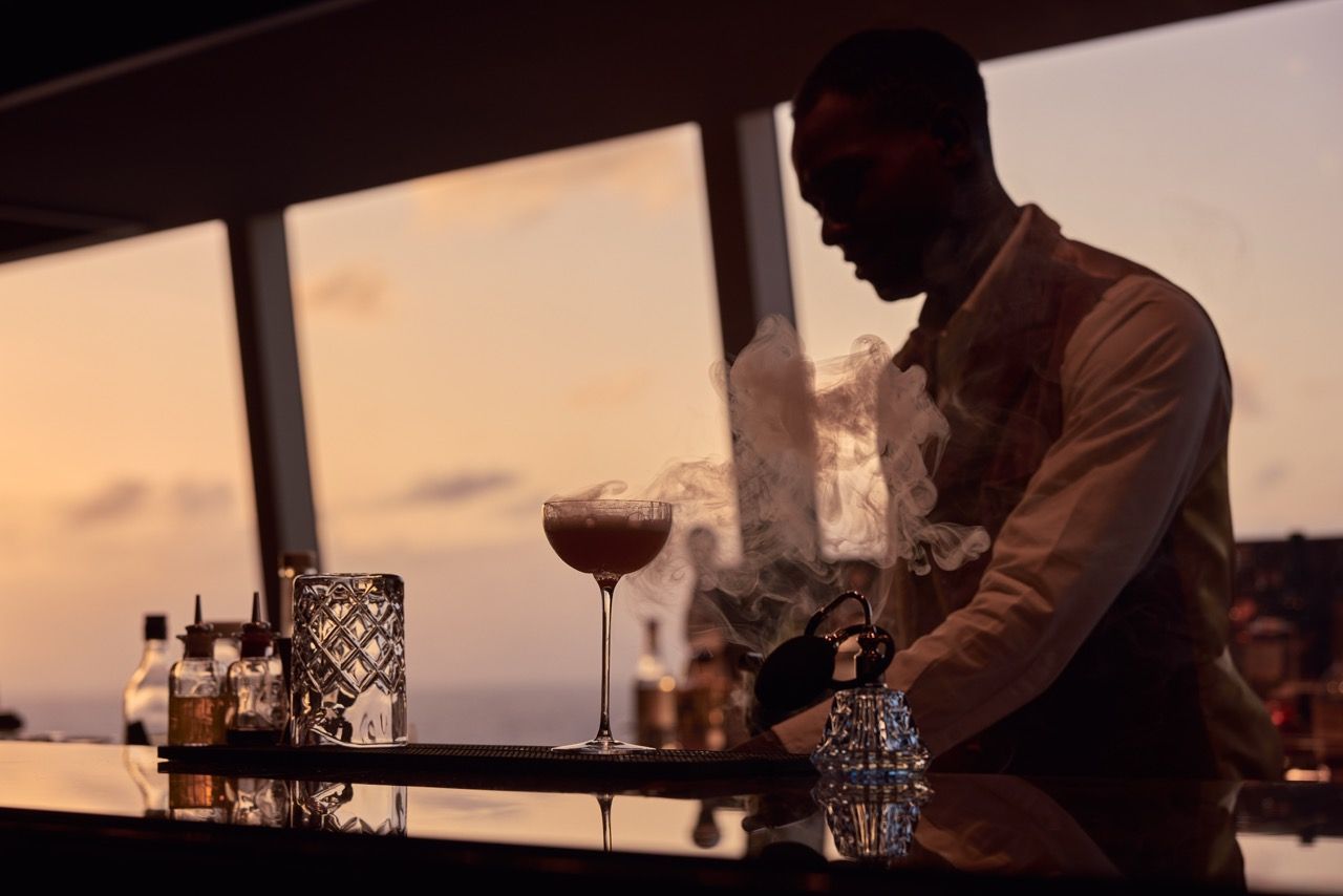 Ein Barkeeper bereitet an einer gl&auml;nzenden Theke vor einem gro&szlig;en Fenster mit Meerblick einen rauchenden Cocktail zu.