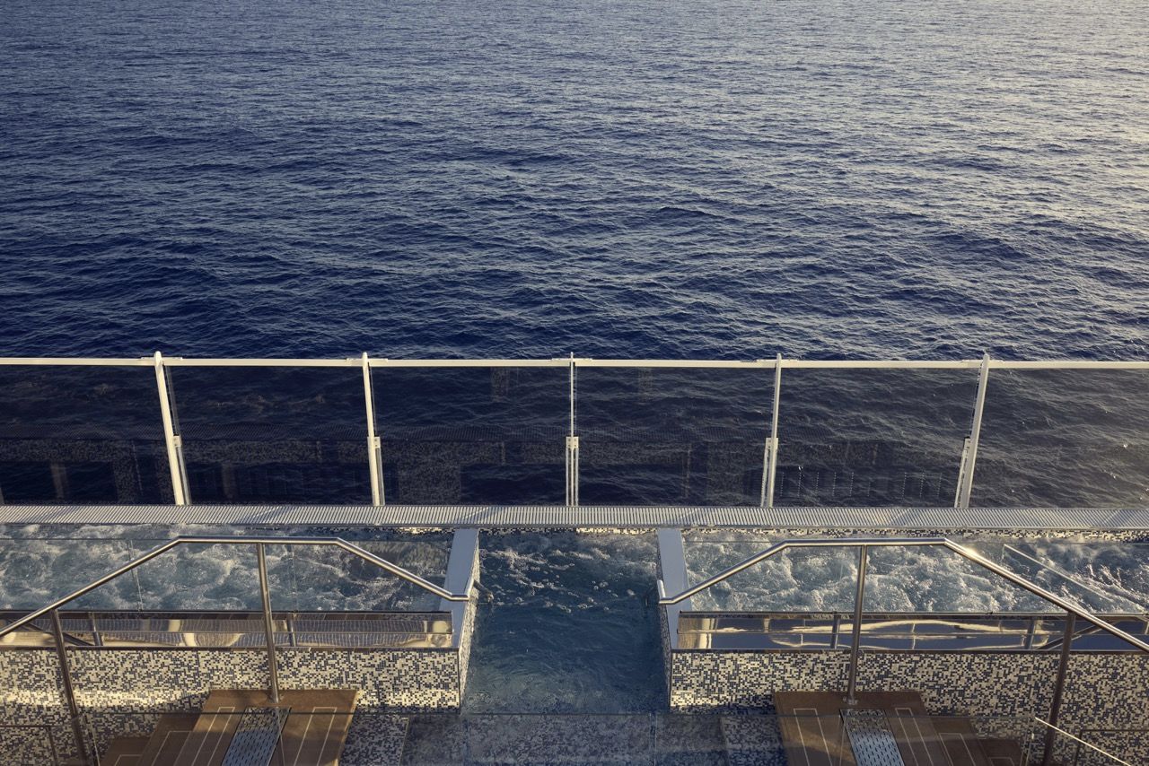 Ein Whirlpool an Bord eines Kreuzfahrtschiffs mit Mosaikfliesen und Handl&auml;ufen aus Edelstahl bietet einen freien Blick auf das tiefblaue Meer durch eine Glasreling.