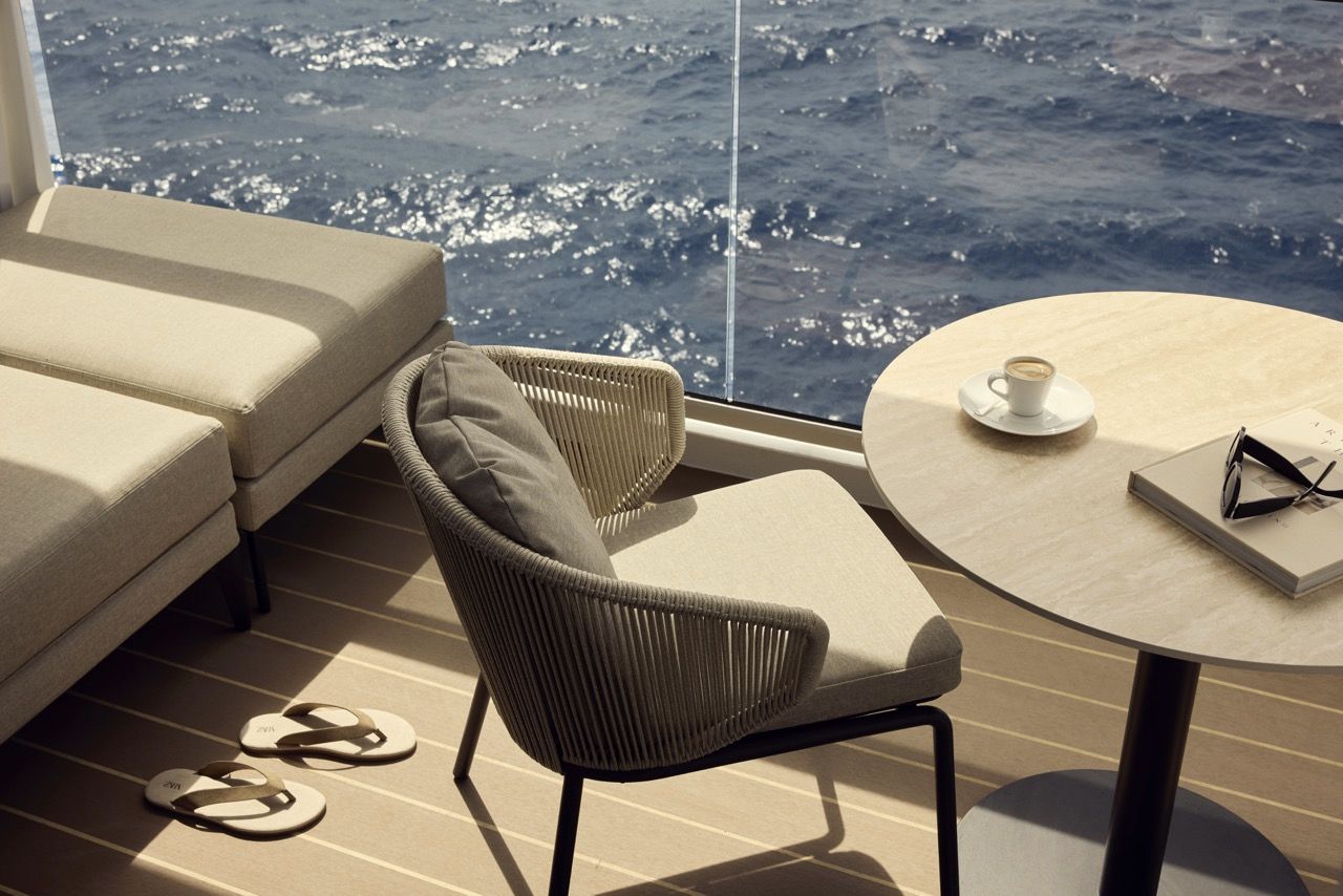 Detailaufnahme einer sonnigen privaten Suite-Terrasse an Bord von Explora Journeys mit hellem Holzboden, einem eleganten Sessel in Seiloptik, einem runden Tisch mit Kaffeetasse und Buch sowie einem Liegestuhl, mit Blick durch eine Glasscheibe auf das wellige, blaue Meer.